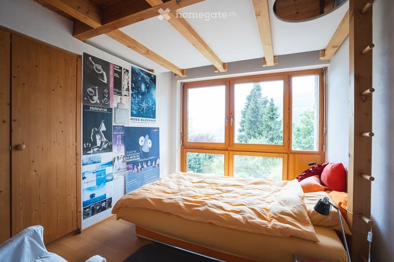Cozy bedroom with wooden beams, large window offering a view, bed with orange bedding, posters on wall, and a small lamp on the nightstand.