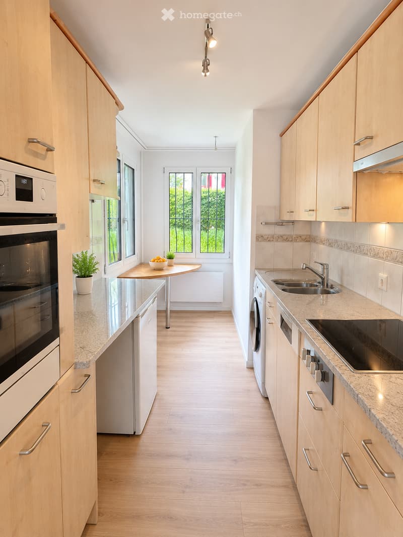 Modern kitchen with light wooden cabinets, granite countertops, stainless steel appliances, and a small round table with a potted plant.