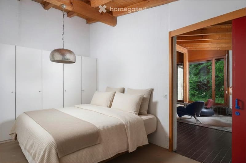 Modern bedroom with a large bed, white pillows, built-in wardrobes, and a pendant light. An open doorway leads to a seating area with a view of greenery.