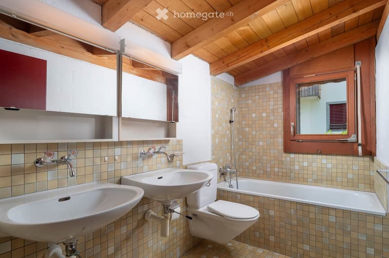 Bathroom with two sinks, a toilet, a bathtub, and a window with wooden frame. The walls are tiled, and there is a wooden ceiling with exposed beams.