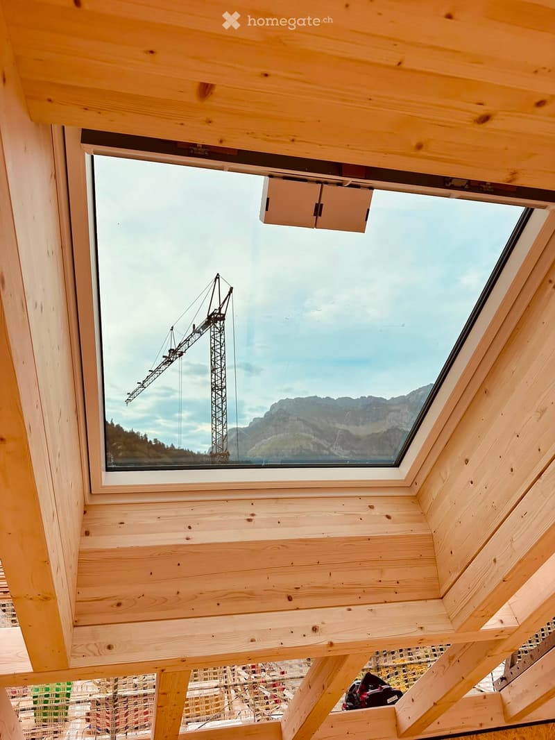 View from inside a wooden structure showing a large skylight window with a view of mountains and a construction crane outside.