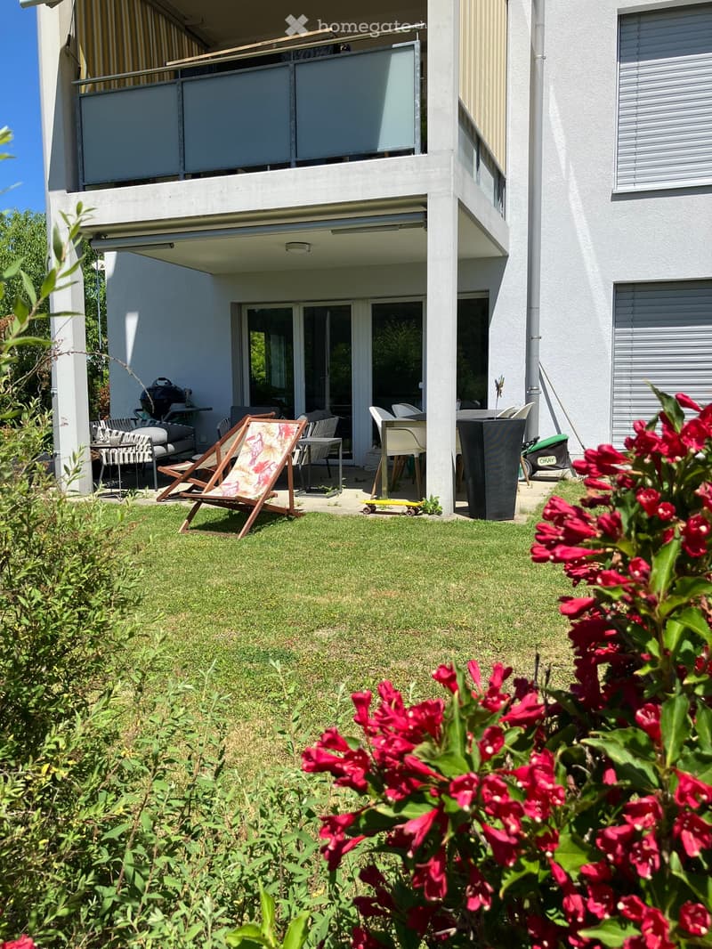 Garden area with a lounge chair, dining table, chairs, and a skateboard near a modern building with balconies.