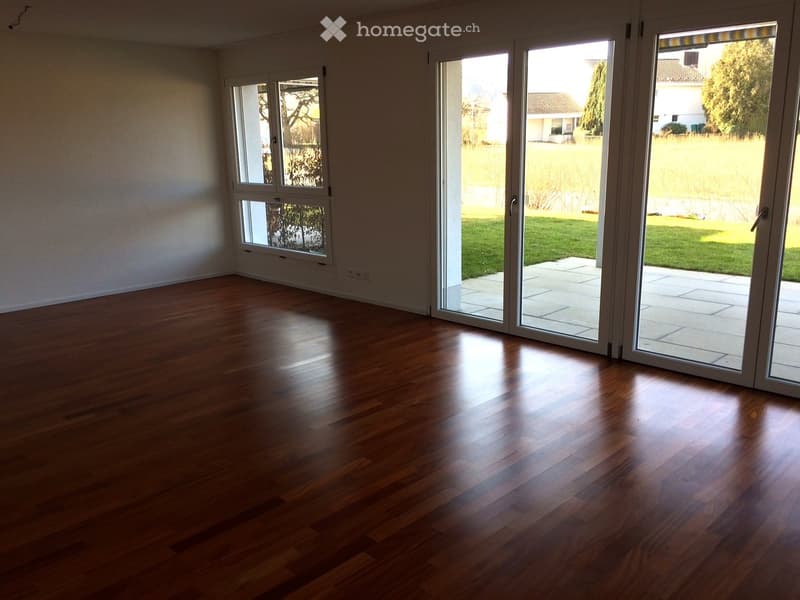 Empty room with wooden flooring, large sliding glass doors leading to a patio and garden, and a window on the left side.