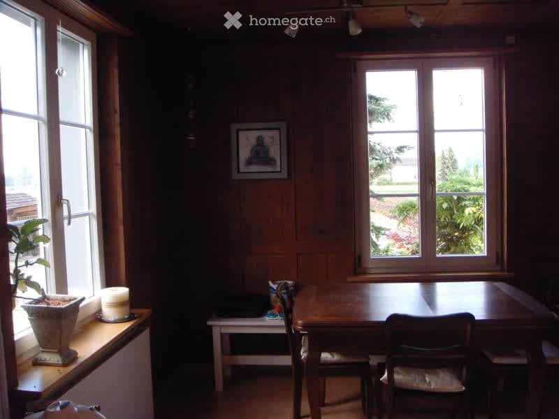 Bright room with wooden walls and large windows, featuring a dining table, chairs, a small bench, a candle, a potted plant, and a picture frame on the wall.