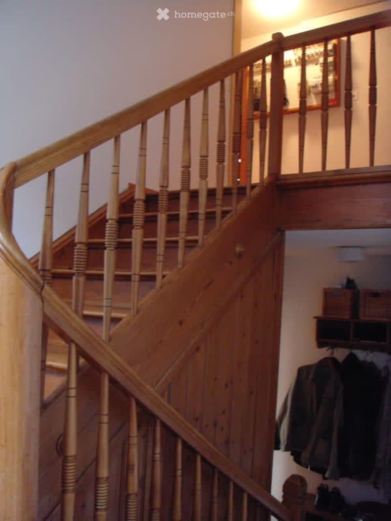 Spiral staircase with wooden railings and handrails, leading to an upper level. The walls are painted in a light color, and there is a small shelf with folded clothes visible on the right side.