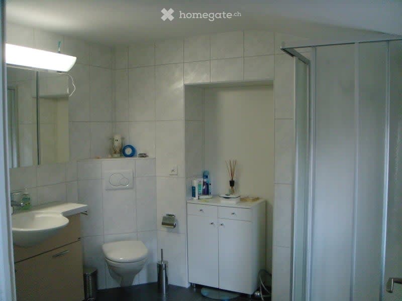 Modern bathroom with white tiles, a sink, toilet, mirror, and a small cabinet. A shower area is visible on the right side.