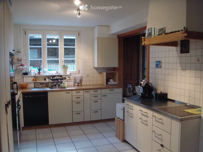 Bright kitchen with white tiled walls and floor, white cabinets, and stainless steel appliances including a dishwasher, microwave, and coffee maker. The countertop has a sink, faucet, and various small items.