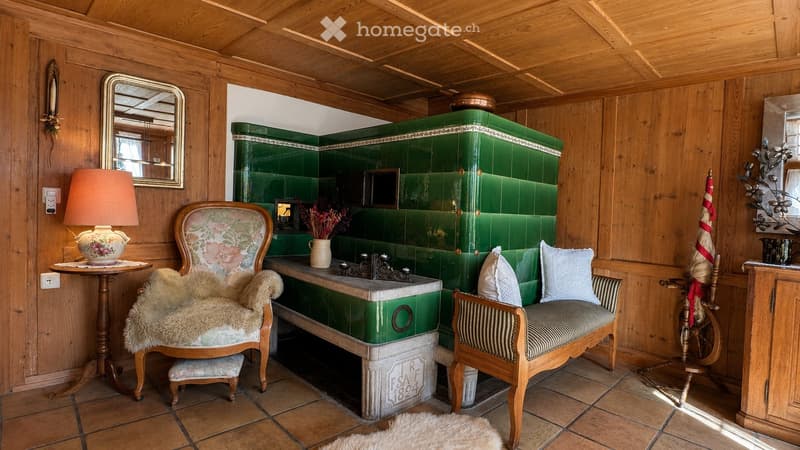 Cozy room with wooden walls and ceiling, green tiled stove, floral-patterned chair, small table with lamp, and decorative items.