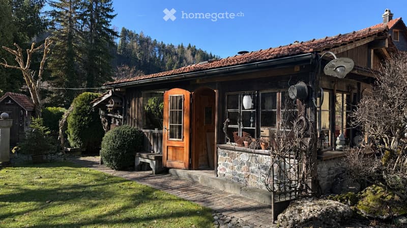 A rustic wooden house with a tiled roof, surrounded by a well-maintained garden with trees, shrubs, and a stone pathway leading to the entrance.