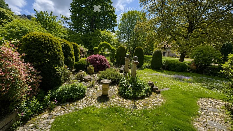 Well-maintained garden with neatly trimmed hedges, flowering shrubs, a stone pathway, and a central stone bench. The garden features a variety of plants and trees, with a lawn and a small fountain in the background.