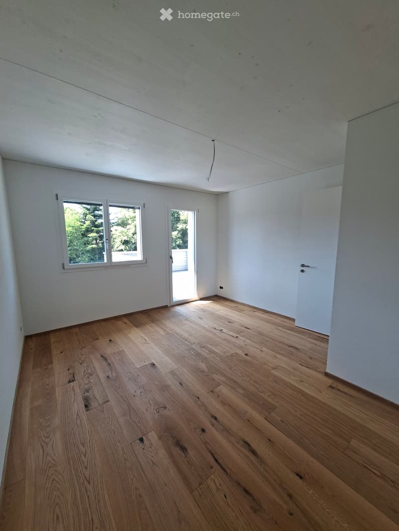 Empty room with wooden flooring, white walls, and a window with a view of greenery. The room has a door leading to another space.