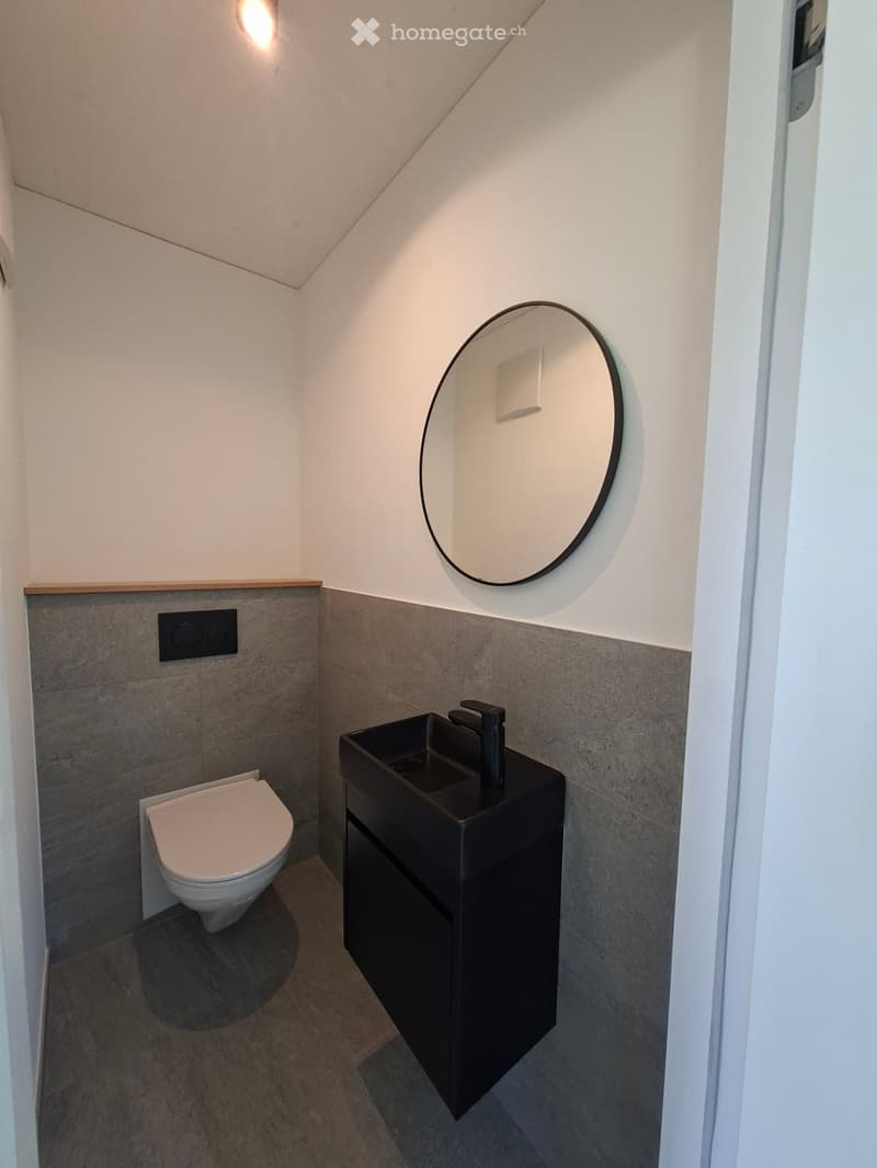 Modern bathroom with a sleek black sink, round mirror, and white toilet, featuring grey tiled walls and a wooden trim.