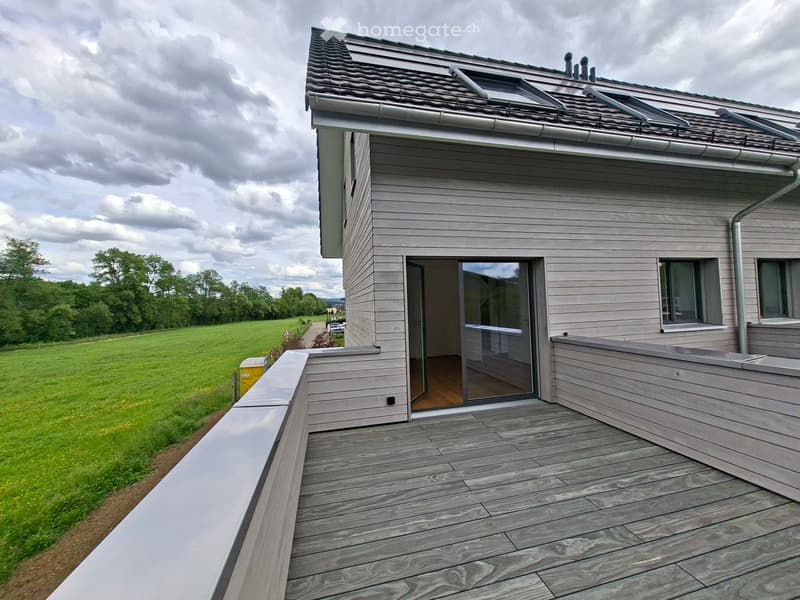 House. Modern two-story house with grey wooden siding, a sloped roof, and a large wooden deck extending from the entrance.