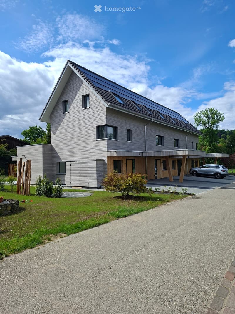 House. Modern two-story house with grey siding, large windows, and a wooden deck area. The house features a sloped roof with solar panels and is surrounded by a well-maintained lawn and gravel driveway.