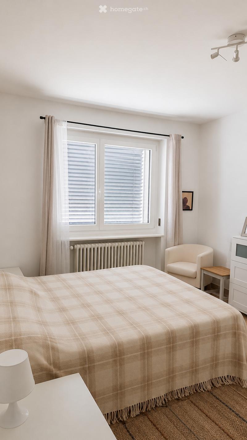 Bright bedroom with a large window, a bed with a plaid blanket, a white armchair, a small wooden side table, a white lamp, and a radiator below the window.