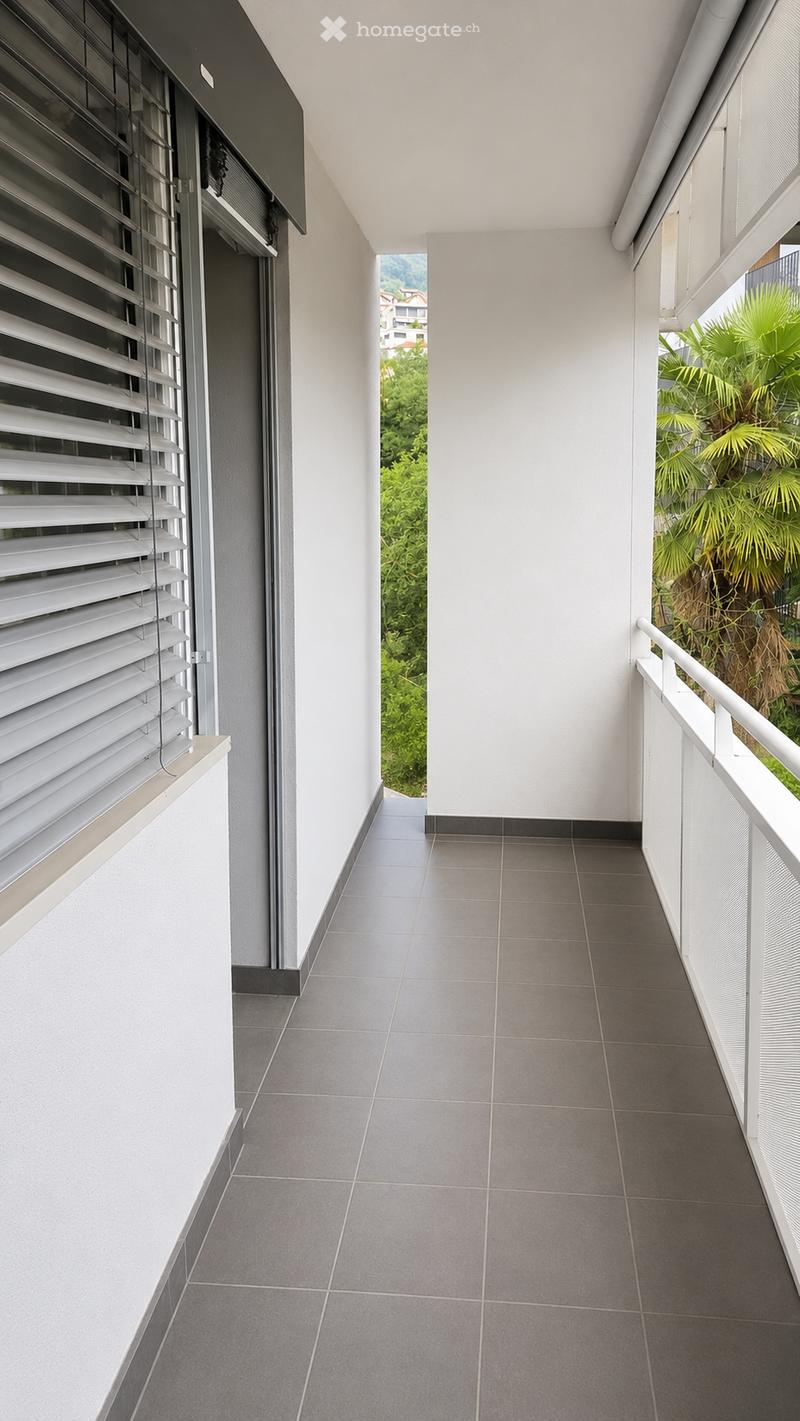 Modern balcony with grey tiled flooring, white walls, and a white metal railing. Features a window with white blinds and a view of greenery and buildings.