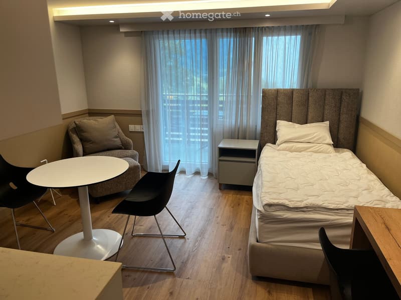 A small bedroom with a single bed, a couch, two chairs, a round table, a desk, and white curtains covering the window that leads to a balcony.