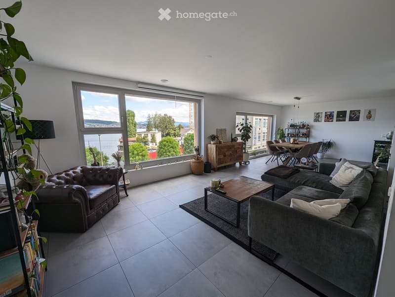 Bright living room with large windows offering a scenic view, featuring a leather sofa, grey sectional sofa, coffee table, dining area with chairs, and decorative plants.