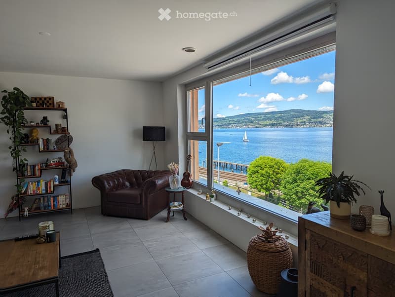 Modern living room with large windows offering a scenic view of a lake and mountains, featuring a brown leather sofa, wooden coffee table, bookshelf, floor lamp, and decorative items.