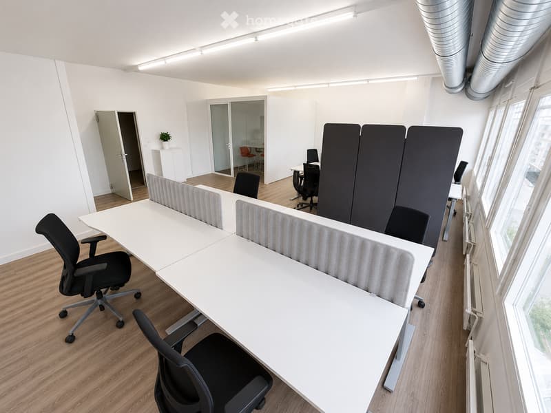 Modern office space with white tables, black chairs, and privacy screens. Large windows provide natural light, and the floor is wooden.