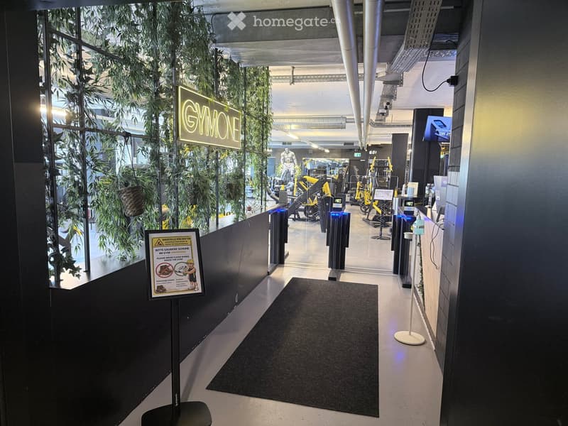 Entrance area of a gym with a sign reading "GYMONE" and visible exercise equipment beyond the glass doors.