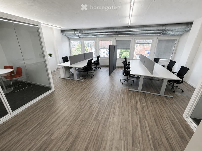 Modern office space with wooden flooring, large windows, white tables, black chairs, and privacy screens.