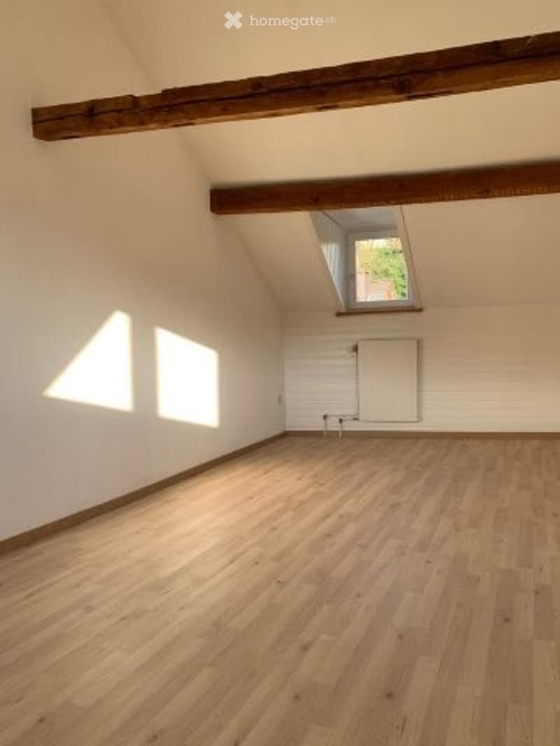 Completely empty room with wooden floor, white walls, exposed wooden beams, and a skylight window.