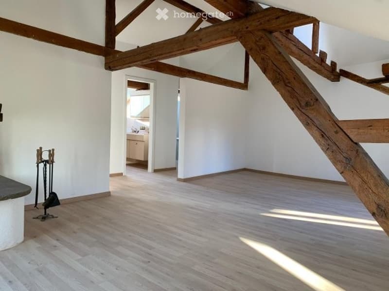 Empty room with wooden floor, white walls, exposed wooden beams, and a small adjacent kitchen area visible through an open doorway.