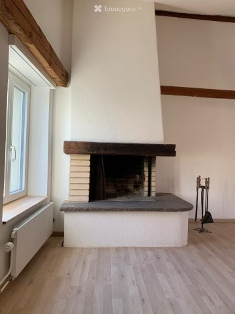 Empty room with wooden flooring, white walls, and a fireplace with a wooden mantel and white surround. A window with white frame and radiator is visible on the left side.