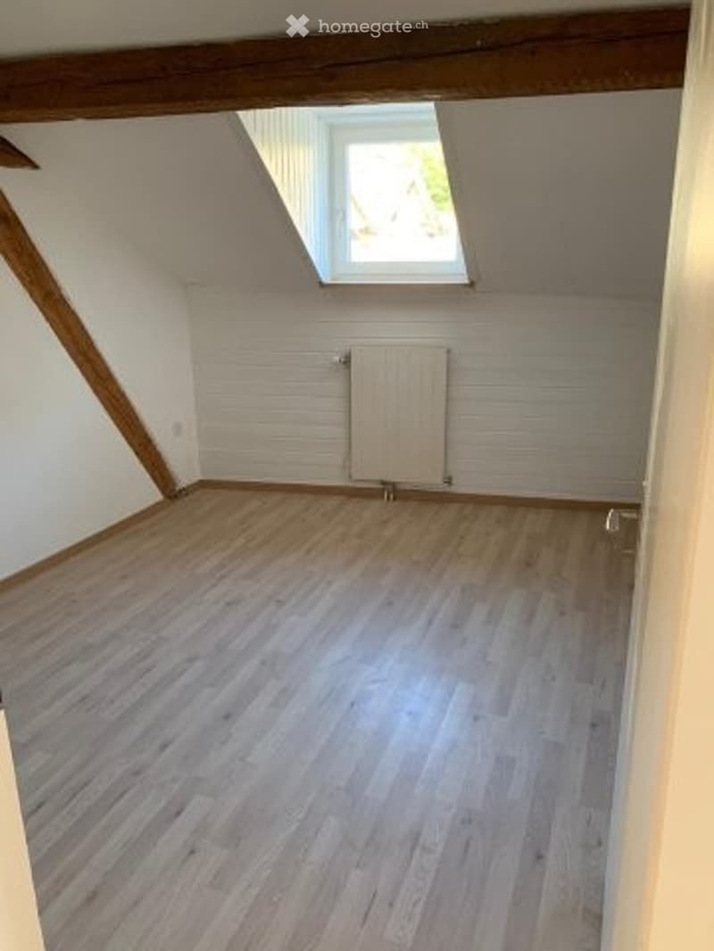 Empty attic room with wooden flooring, white walls, exposed wooden beams, and a skylight window.