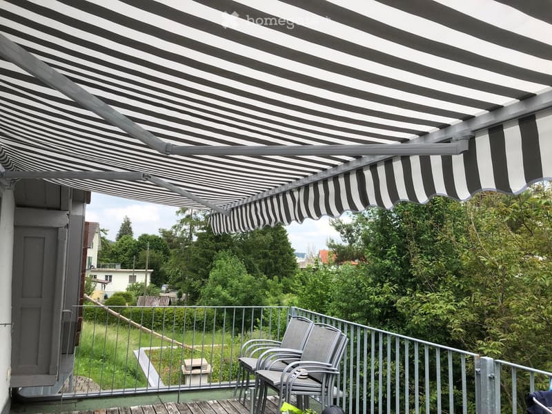 Balcony with a retractable awning, metal railing, and two grey chairs.
