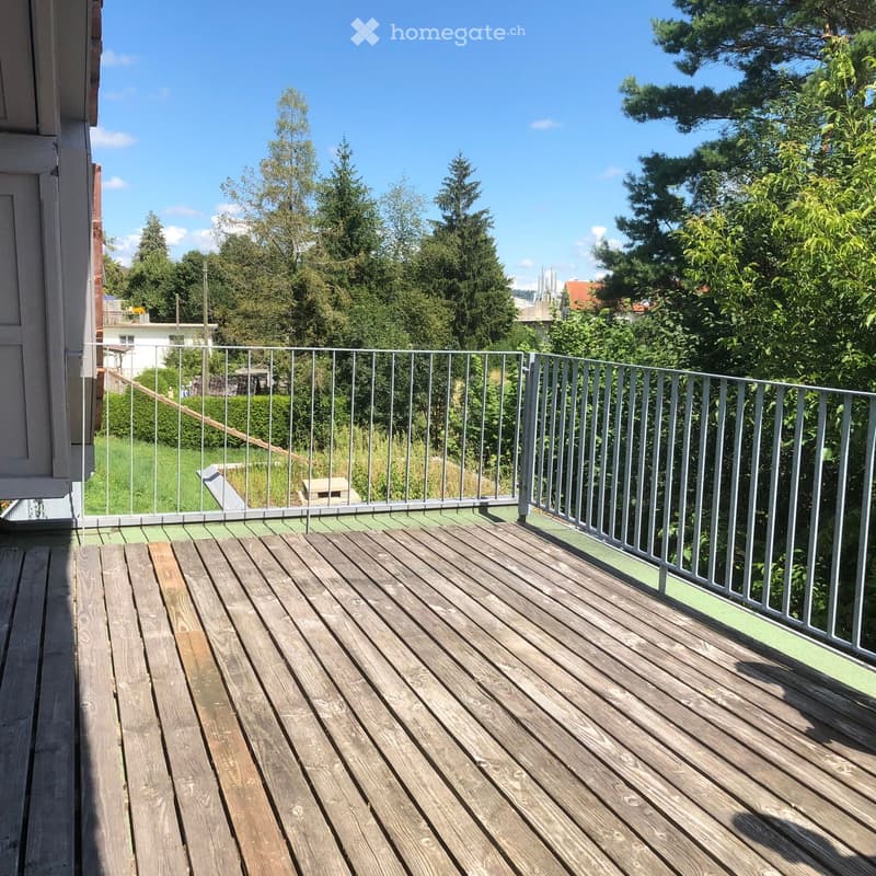 Wooden deck with metal railing, overlooking a green, tree-filled area with a clear blue sky.