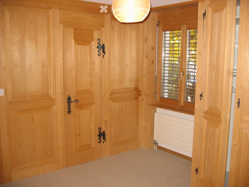 Empty hallway with wooden paneled walls, a closed wooden door, and a window with blinds. The floor is carpeted, and there is a radiator under the window.