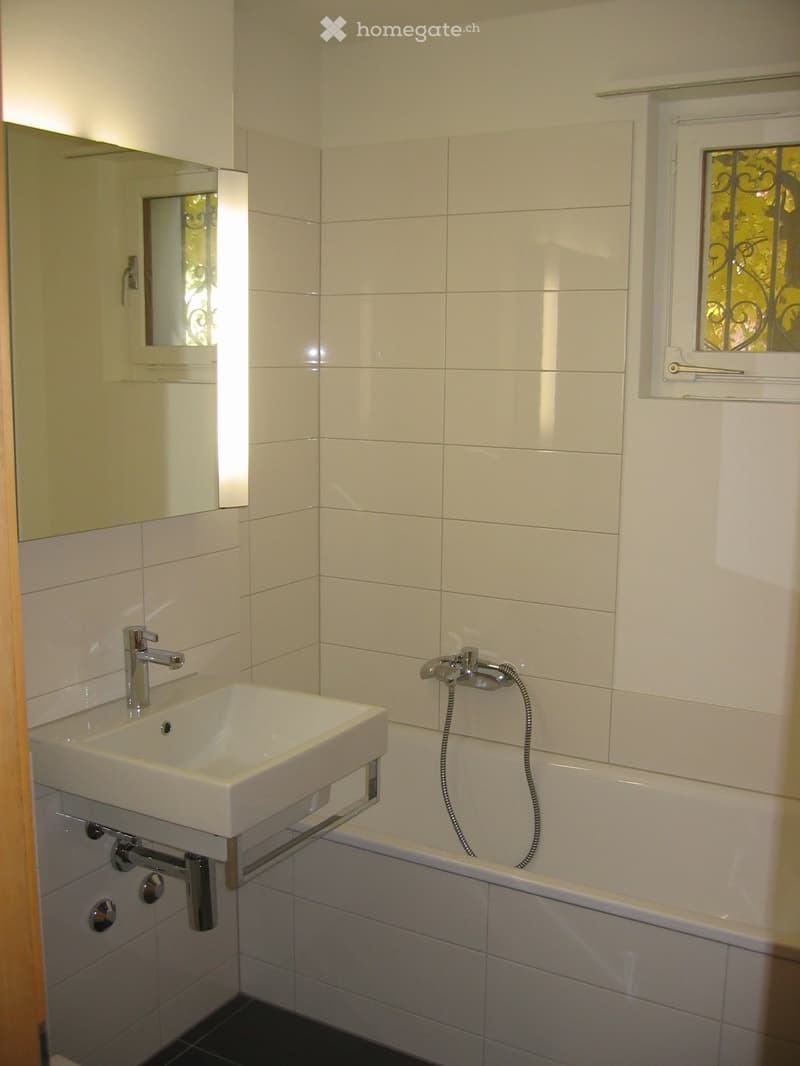 Modern bathroom with white tiled walls, a white sink with chrome faucet, a bathtub with a handheld shower hose, and a mirrored cabinet.