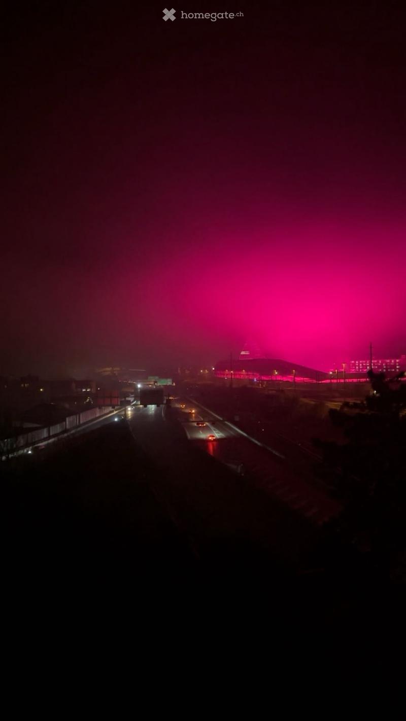 Nighttime view of a highway with traffic, distant illuminated building, and pink sky.