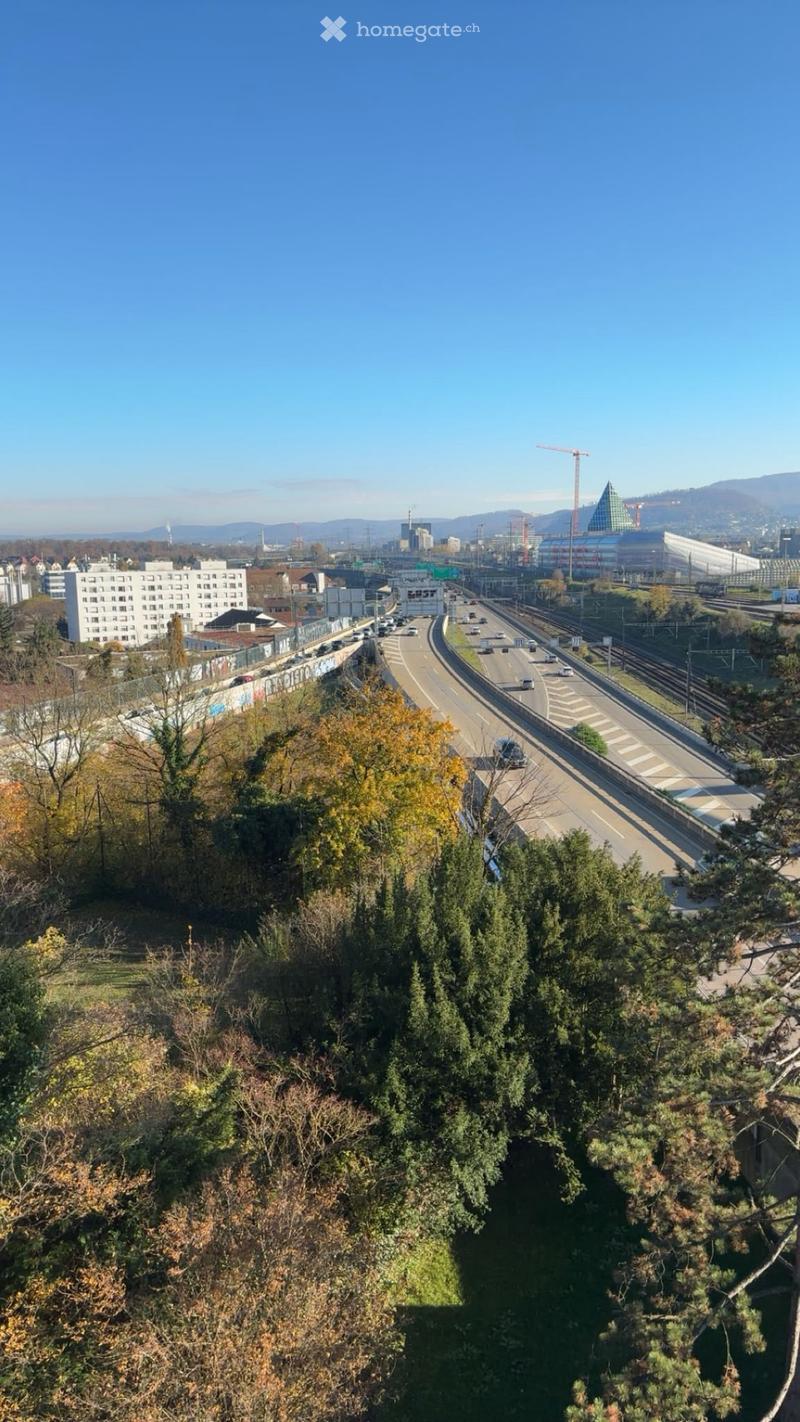 A panoramic view of a cityscape with a multi-lane highway, railway tracks, and surrounding buildings under a clear blue sky.