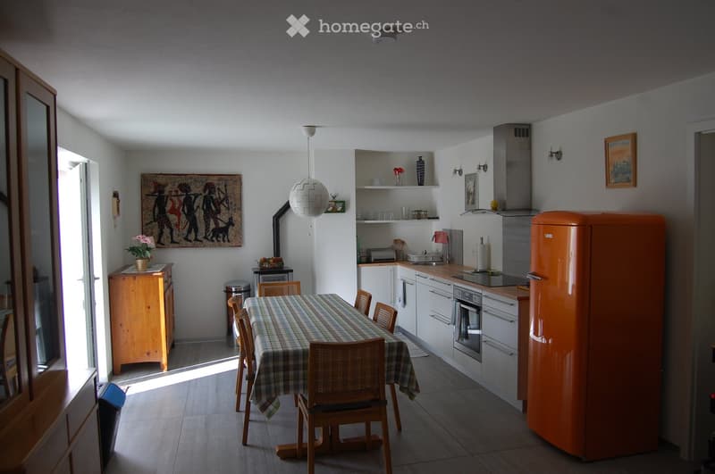 Bright kitchen with white cabinets, an orange refrigerator, a dining table with chairs, and a pendant light. Features a wooden sideboard, a painting, and a vase on a shelf.