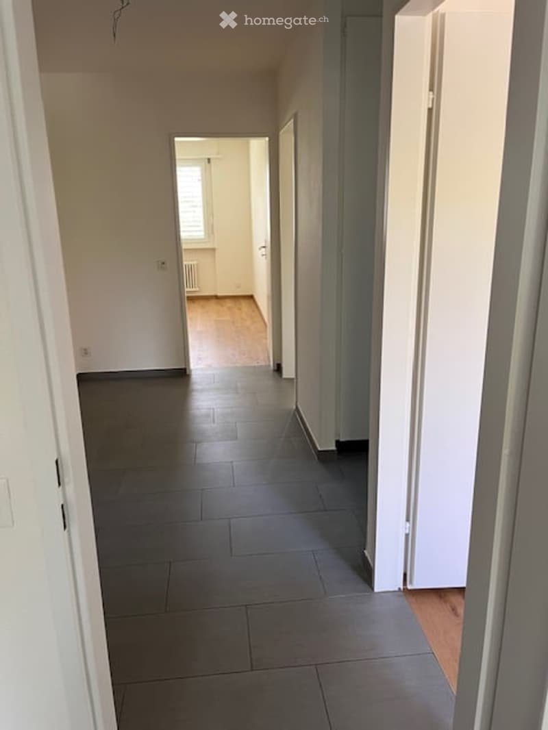 Empty hallway with grey tiled floor, white walls, and a visible room at the end with wooden flooring and a window.