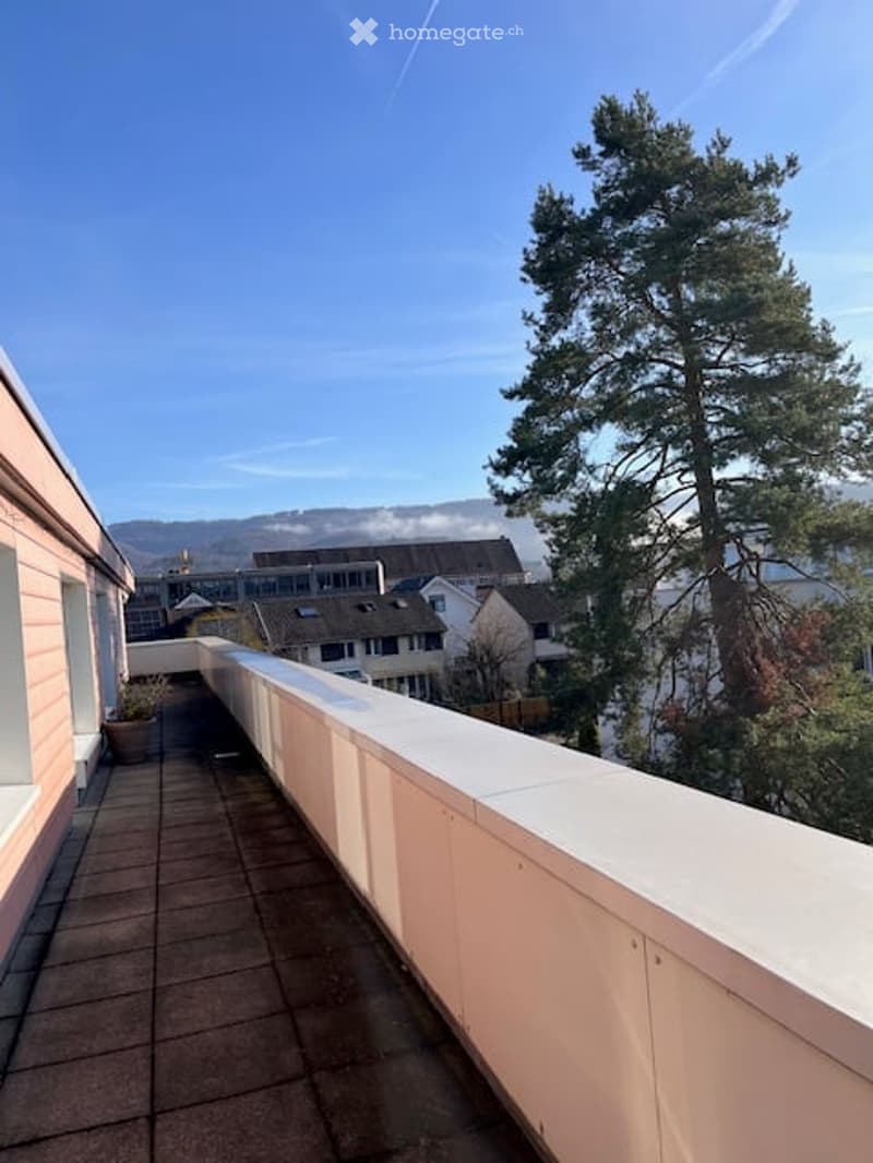 Balcony with white railing, tiled floor, overlooking a residential area with trees and distant mountains.