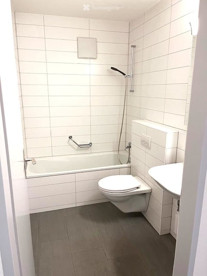 Modern bathroom with white tiled walls and floor, featuring a bathtub, toilet, sink, and a showerhead with a handrail.