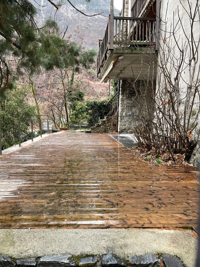 Wooden deck leading to a stone house with a balcony, surrounded by trees and a mountainous landscape.