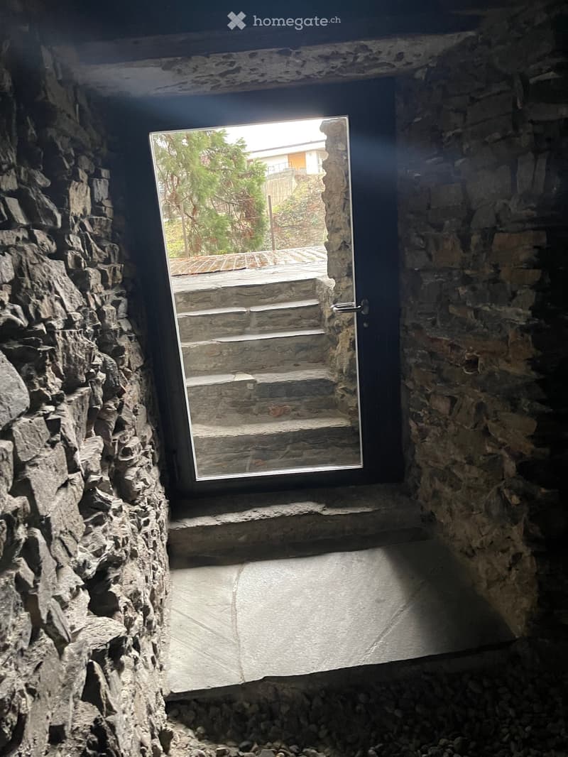 A narrow stone-walled corridor with a large window opening to an outdoor staircase and garden area.