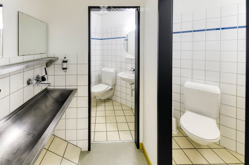 Modern bathroom with white tiled walls and floors, featuring a sink with a faucet, a mirror, and a toilet. The design includes a sleek, metallic sink basin and a clean, minimalist aesthetic.