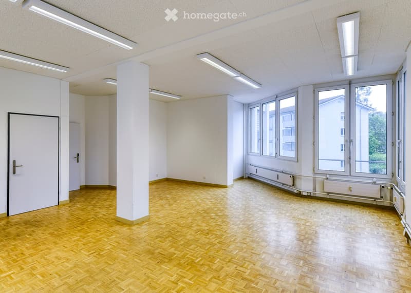 Empty room with wooden herringbone flooring, white walls, large windows, and a central column. No furniture or equipment present.