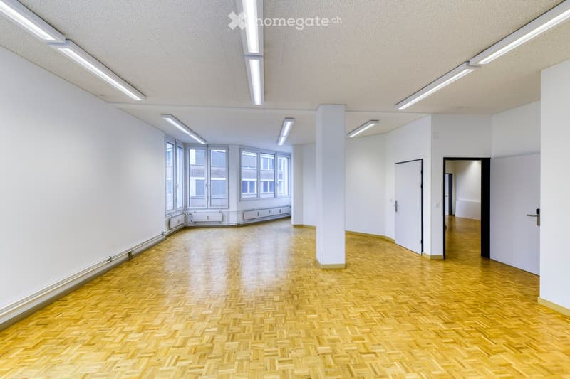 Empty room with wooden herringbone flooring, white walls, large windows, and recessed lighting. No furniture or equipment present.