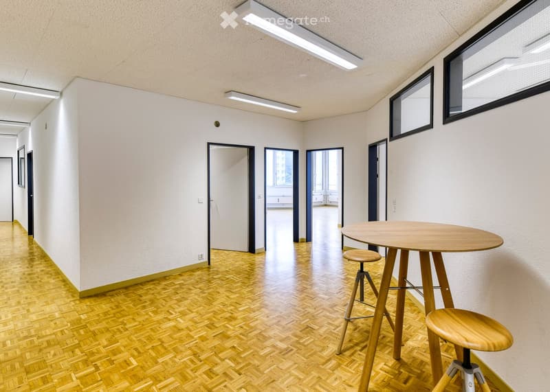A modern hallway with wooden herringbone flooring, white walls, and several doors leading to other rooms. A small round wooden table with two matching stools is placed near the wall.
