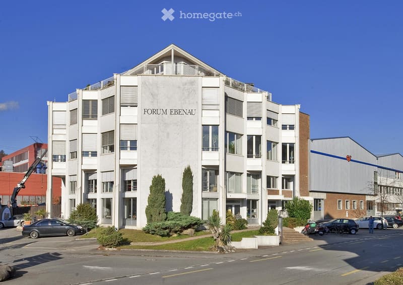 Modern multi-story building with white facade, large windows, and balconies. The building is labeled "FORUM EBENAU" and is surrounded by greenery and parked cars.