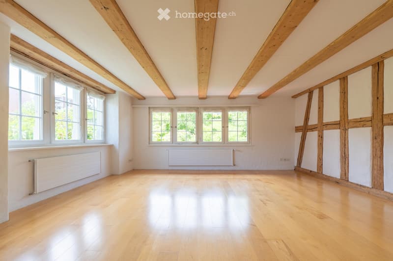 Empty room with wooden flooring, exposed wooden beams on the ceiling, and multiple windows allowing natural light to enter.
