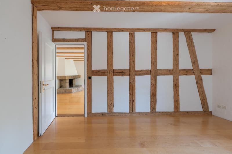 Empty room with wooden floor, white walls, exposed wooden beams, and an open doorway leading to another room with a fireplace.