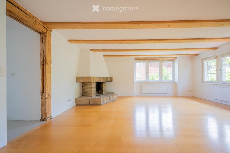 Empty room with wooden floor, exposed wooden beams, and a brick fireplace with a white chimney. Large windows provide natural light.